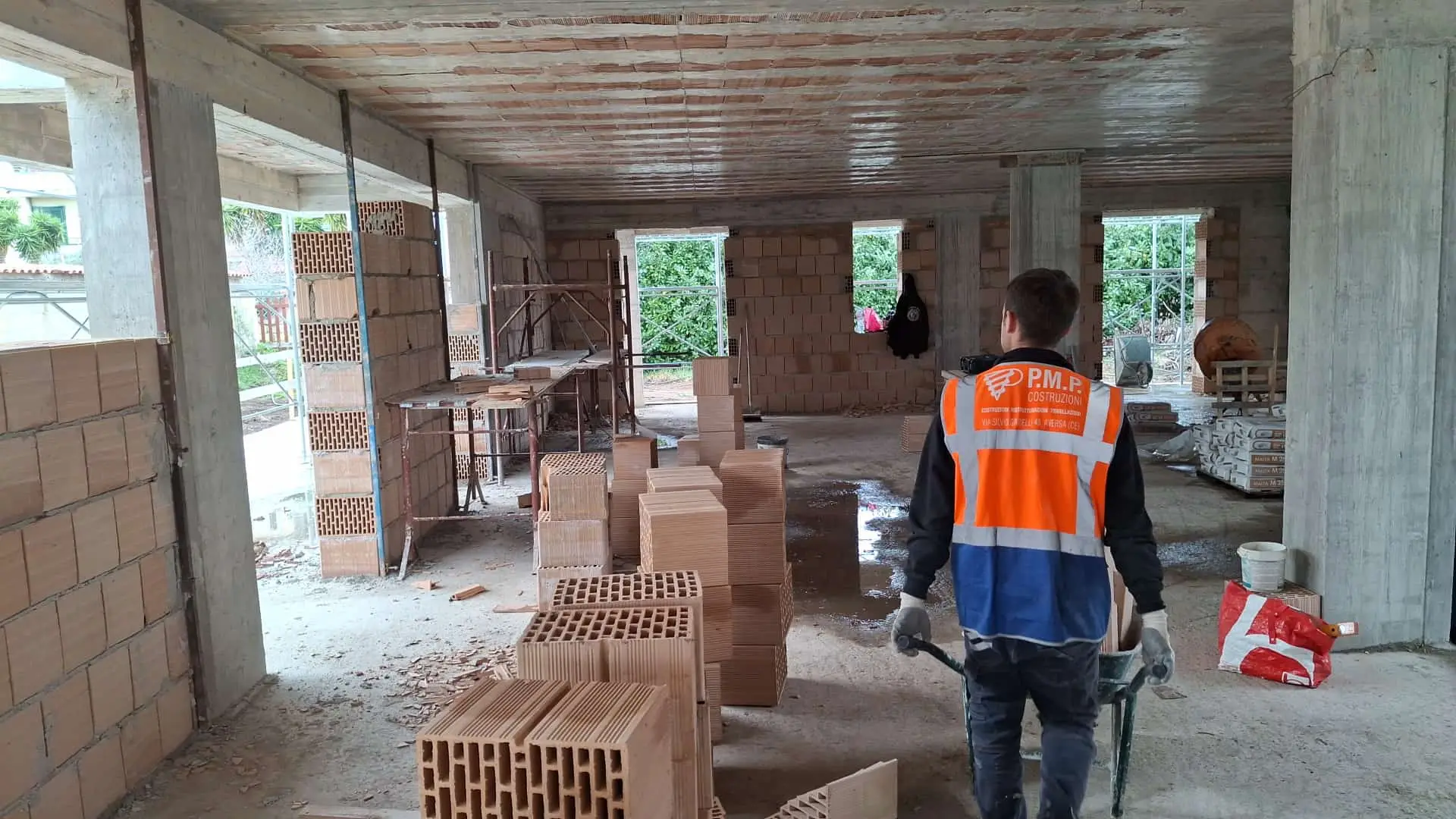 Persona con gilet arancione P.M.P. in un edificio in muratura, con cataste di mattoni.