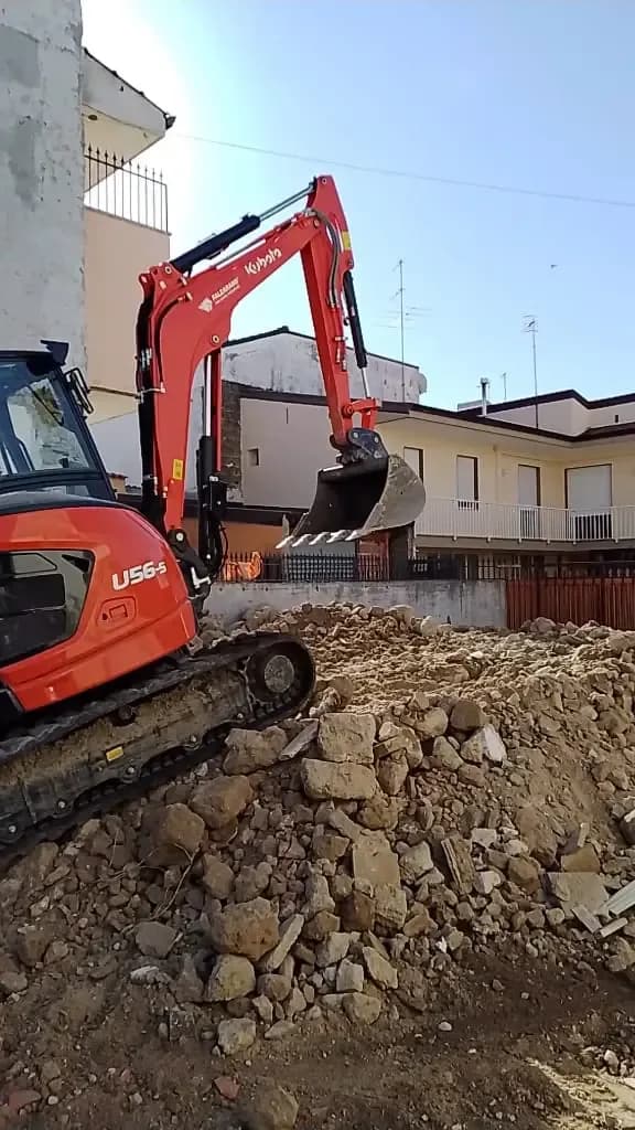 Miniescavatore cingolato al lavoro su un cumulo di terreno di risulta in un cantiere urbano.