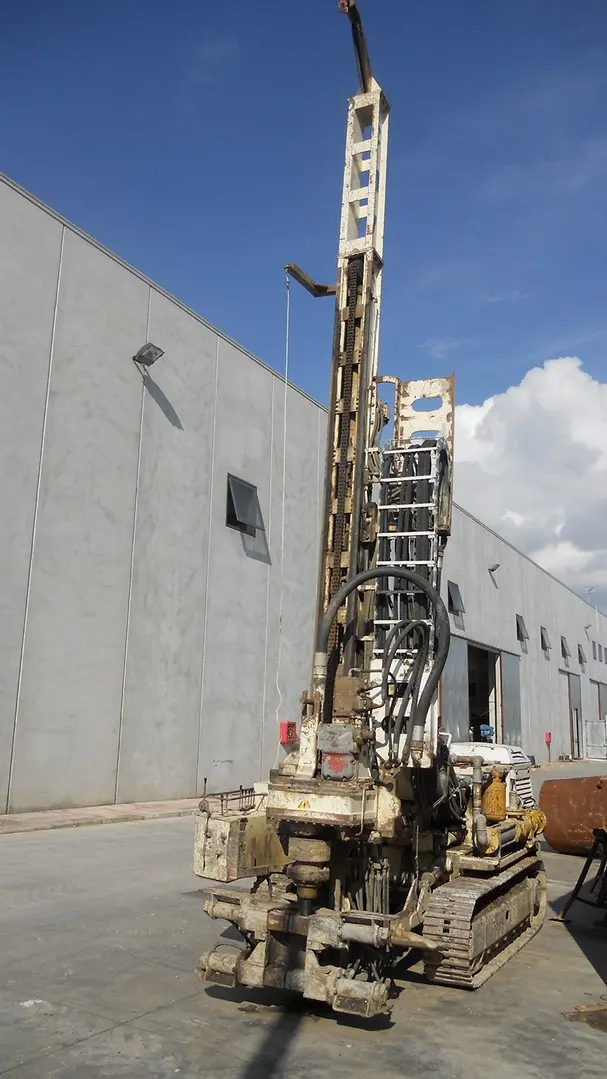 Sonda cingolata con torre di perforazione verticale, fotografata davanti a un capannone.