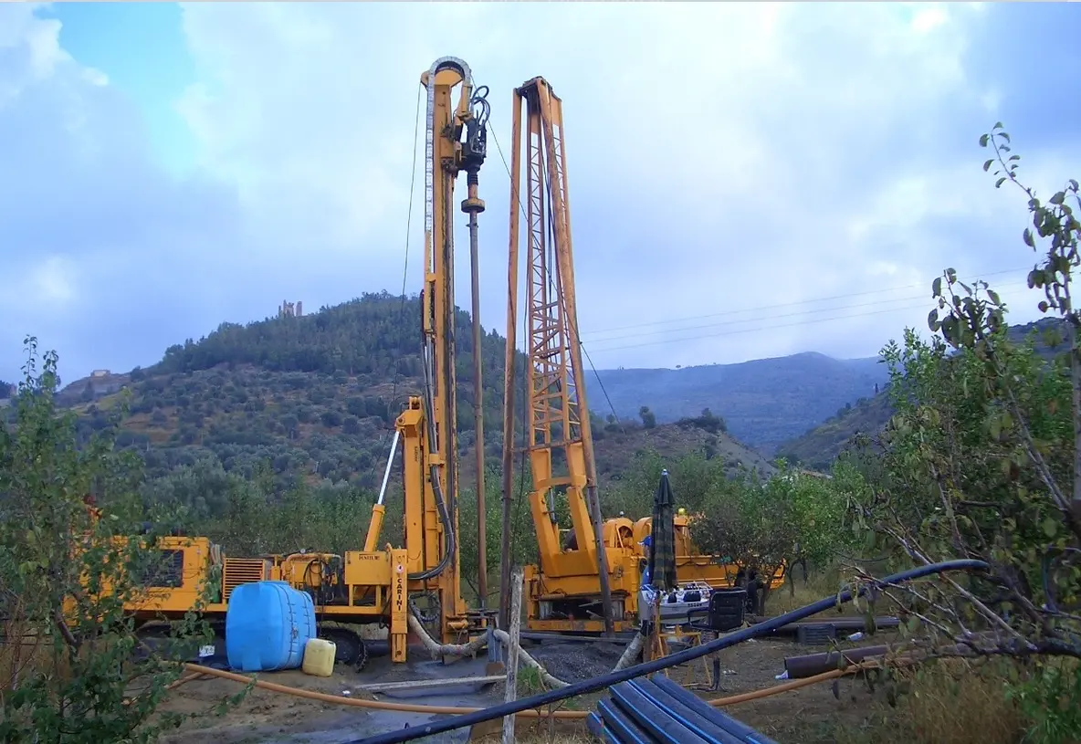 Due sonde di perforazione in un cantiere collinare, con tubazioni e attrezzature per un pozzo.